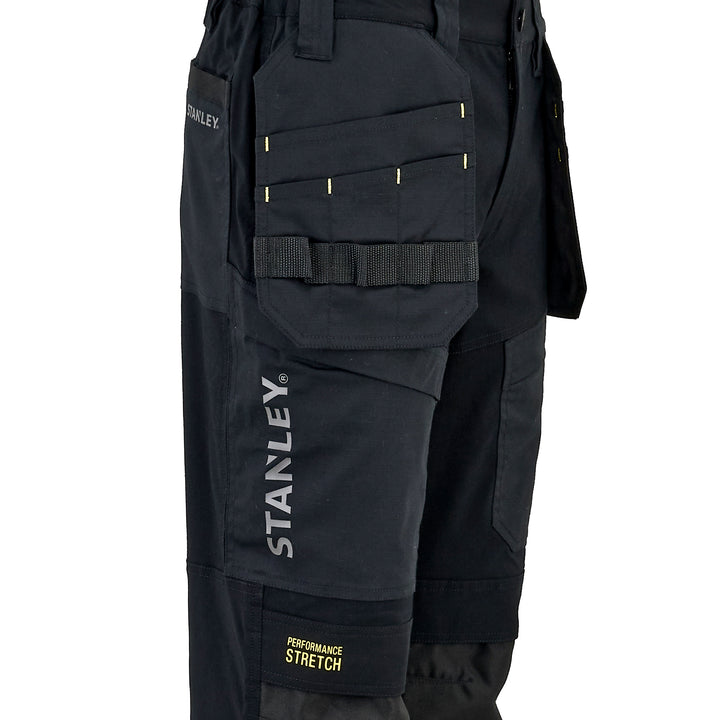 Close-up of black Stanley work pants showing the thigh pocket with yellow reflective stitching, elastic band, and vertical white STANLEY branding, plus a small yellow Performance Stretch label near the hem.