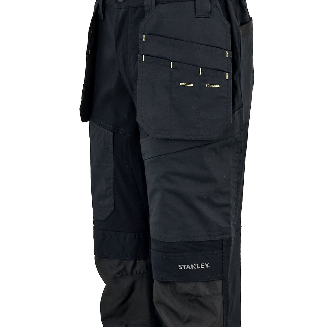 Close-up of black work pants showing the right leg with multiple utility pockets, including a large front pocket with gold-tone zipper details and a lower cargo pocket with the white STANLEY logo.
