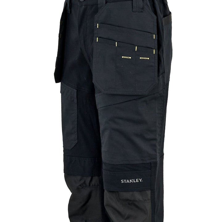 Close-up of black work pants showing the right leg with multiple utility pockets, including a large front pocket with gold-tone zipper details and a lower cargo pocket with the white STANLEY logo.