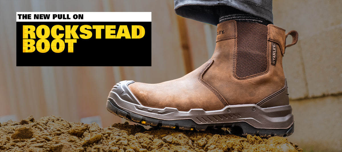A brown leather work boot with a pull tab, shown being worn with jeans on dirt, with a black and yellow Rocksteady Boot promotional overlay on the left.