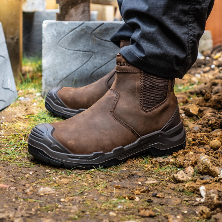 A pair of brown leather work boots with black rubber soles, shown being worn outdoors on a dirt and gravel surface next to a stone.