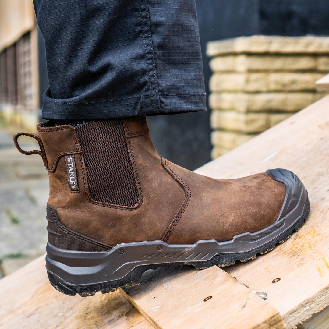 A brown leather ankle work boot with a black rubber toe cap and sole, shown being worn on a wooden platform in an outdoor setting. The brand name 'STANLEY' is visible on the side of the heel.