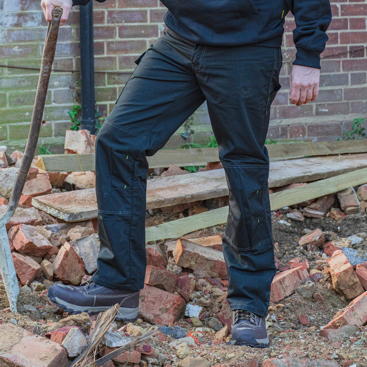 Person wearing dark blue work pants and boots on a construction site with bricks and tools.