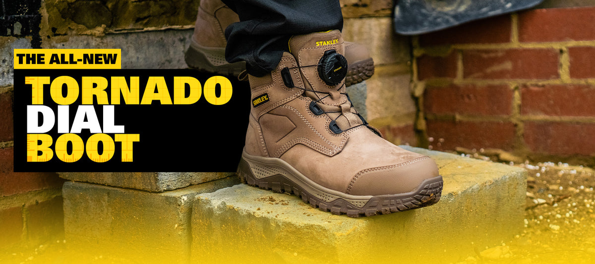A tan work boot with a dial lace closure and black branding, shown worn with dark trousers on stone steps against a brick wall. Large promotional text on the left reads 'THE ALL-NEW TORNADO DIAL BOOT'.