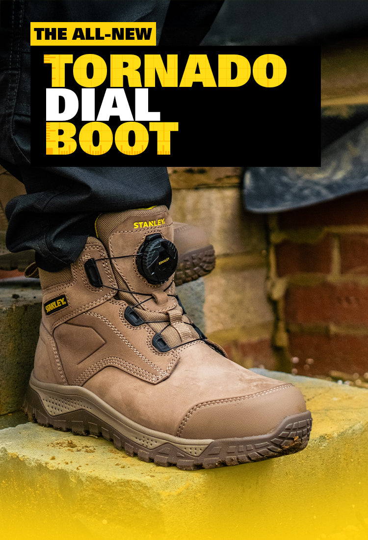 A tan Stanley Tornado Dial Boot work shoe shown on a person's foot, with a black and yellow promotional text banner at the top reading 'THE ALL-NEW TORNADO DIAL BOOT'. The background includes a brick wall and a yellow surface.