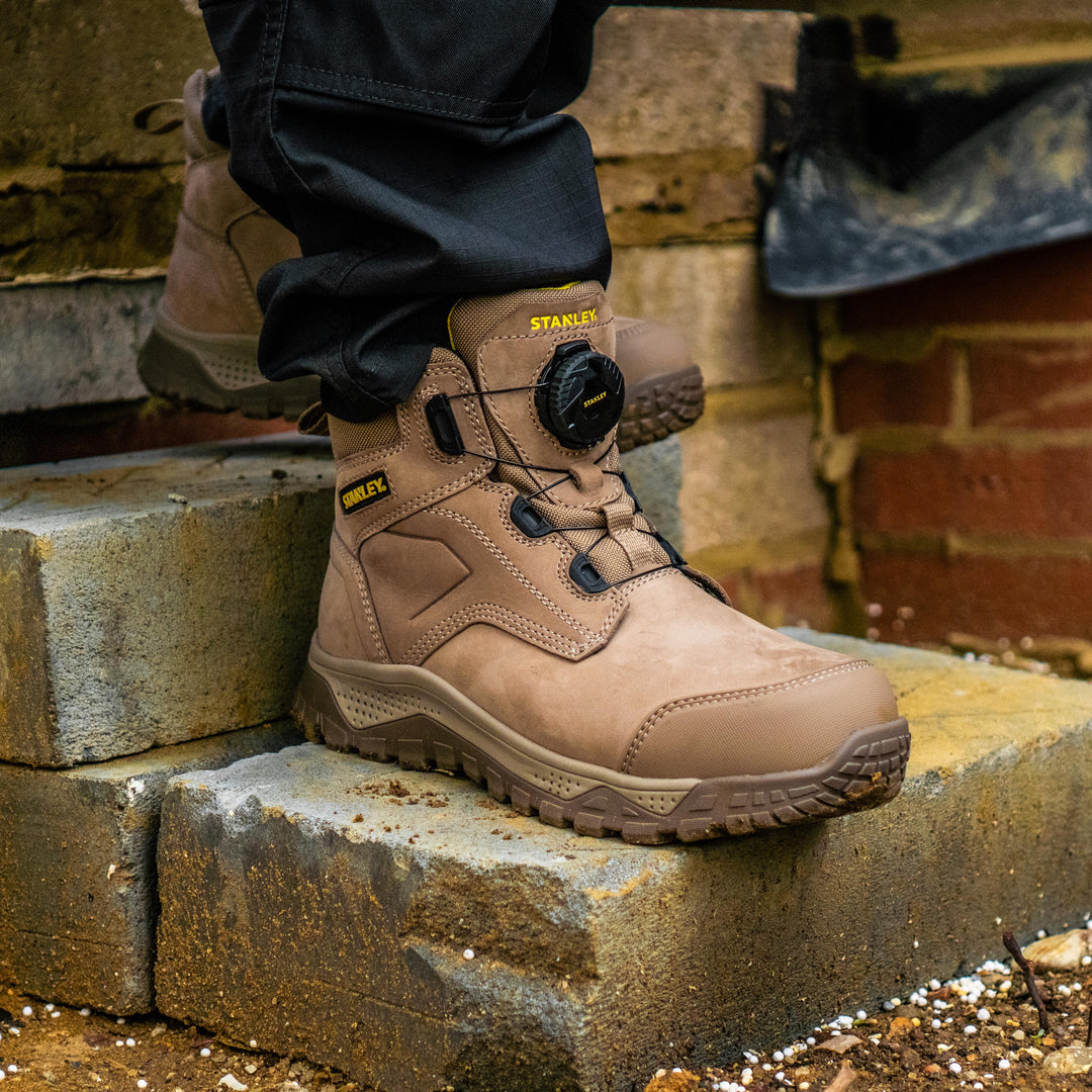 A person wearing a tan Stanley work boot with a black dial closure, standing on stone steps outdoors in front of a brick wall.