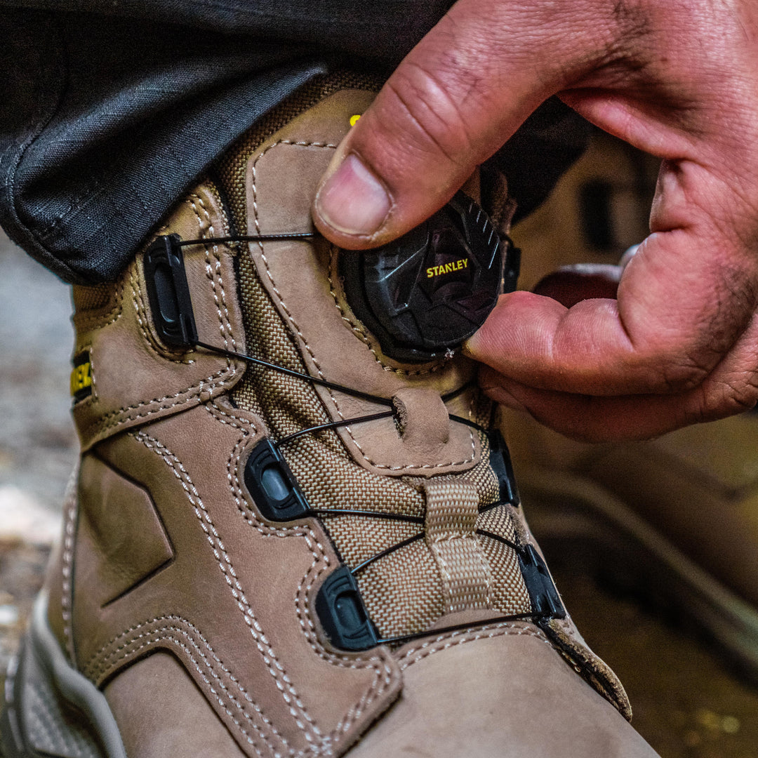 Close-up of a tan work boot with a black dial lace closure being adjusted by a person's hand. The boot has tan suede and canvas panels with black laces and black plastic eyelets.